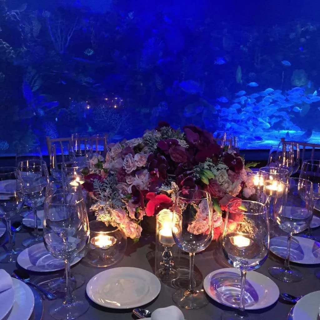 Mesa decorada con arreglos florales frente a un acuario iluminado en tonos azules.