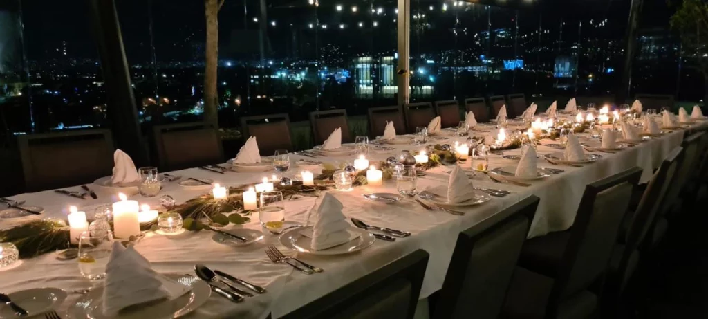 Mesa alargada decorada con velas y servilletas blancas en un evento nocturno con vista a la ciudad.